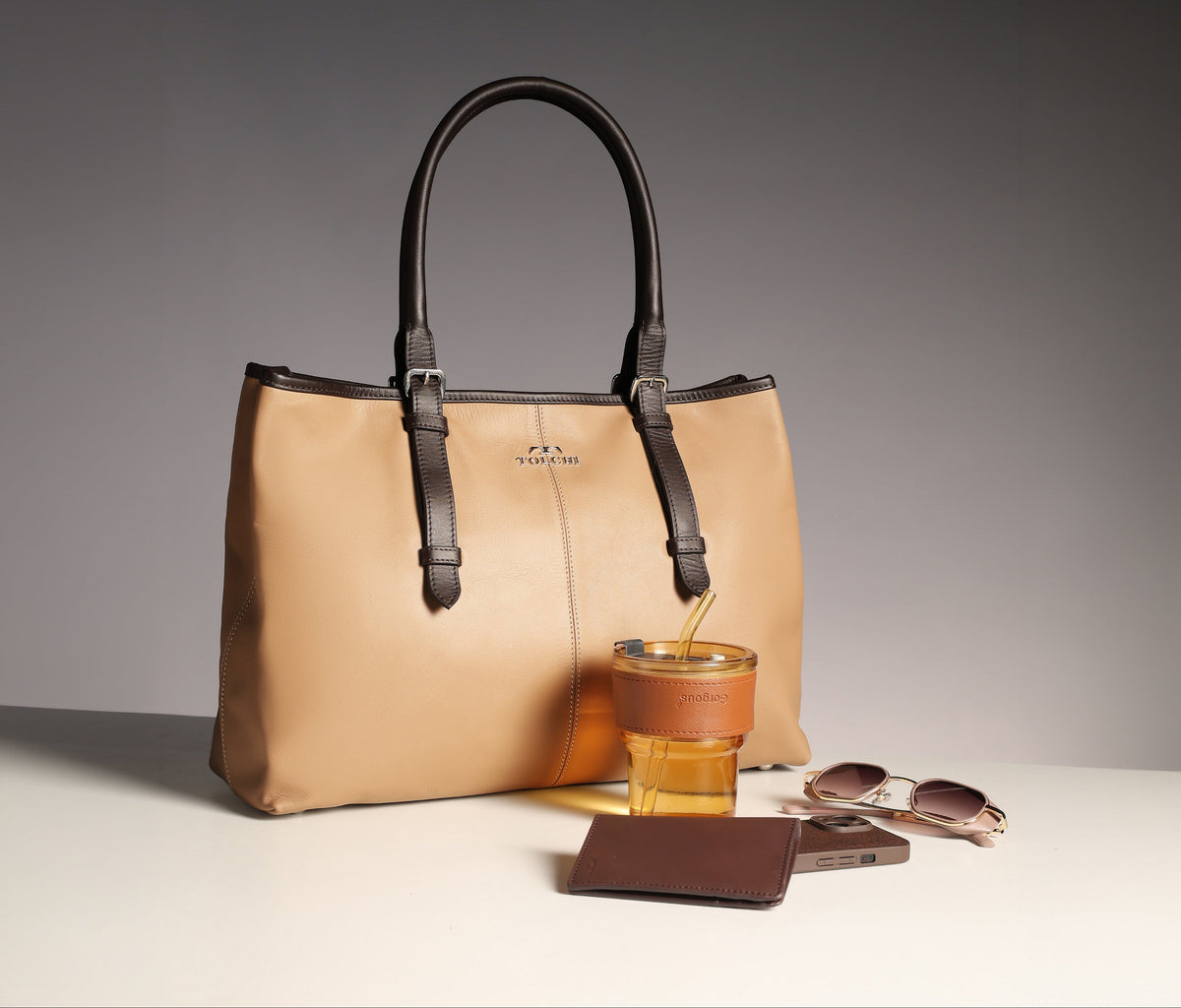 A beige leather tote bag with dark brown handles and trim, styled on a white surface with a glass of iced coffee, sunglasses, a wallet, and a phone against a dark gray gradient background.